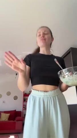 The video features a young woman standing indoors, holding a glass bowl containing a salad. She is fair-skinned with her hair pulled back into a ponytail. She wears a black crop top and loose, light blue pants. She is looking directly at the camera and appears to be expressing disapproval or dislike for the salad. She points to the bowl as if emphasizing her feelings about its contents. A silver fork is also visible in the bowl. 

The background shows a simple living room setting, with a red couch partially visible on the left, and a dark-colored wardrobe on the right. There's also a small red bookshelf with various items on it. The walls are white, and sunlight streams into the room.