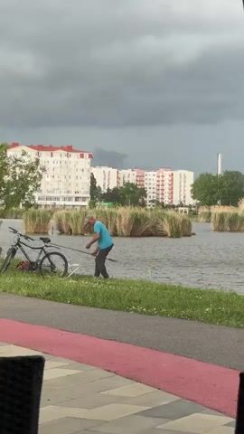 The video shows a man fishing in a lake on a cloudy day. The man is wearing a blue shirt and dark pants. He is standing on the grassy bank of the lake, holding a fishing rod. There is a bicycle leaning against a tree next to him. In the background, there are several apartment buildings and some clumps of tall grass growing in the lake. The camera is positioned from inside a building or under a canopy, showing part of a table and chairs in the foreground. A red path runs along the edge of the lake.