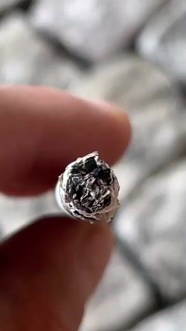 The video shows a close-up of a cigarette being held between two fingers. The cigarette is lit and burning, with ash at the tip. The background appears to be a rough, textured surface, possibly stone. The lighting is natural and the focus is sharp on the cigarette and fingers. The video is brief, lasting only a few seconds, and shows the cigarette being held steady and then slightly rotated.