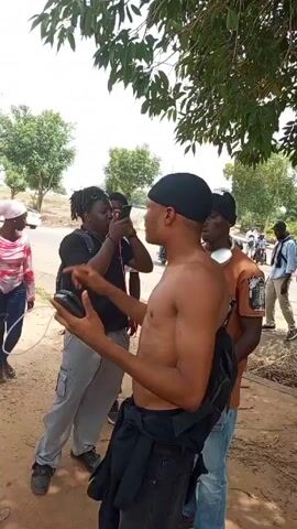 Here is a concise description of the video:

The video appears to capture a scene involving a group of people, possibly related to a protest or demonstration. There are individuals holding signs or banners that read "FREE OUR PRESIDO." Some people are wearing casual clothes, and there is a person with a camera and recording equipment. They are positioned on a dirt road or roadside, with a street and passing cars in the background. The mood seems intent, suggesting a serious atmosphere related to the cause they are supporting.
