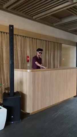A person stands behind a wooden counter, wearing a maroon shirt and sunglasses. To the left of the counter is a tall, black speaker with a smaller speaker on top. The background features a bamboo-like wall covering and a wooden ceiling.