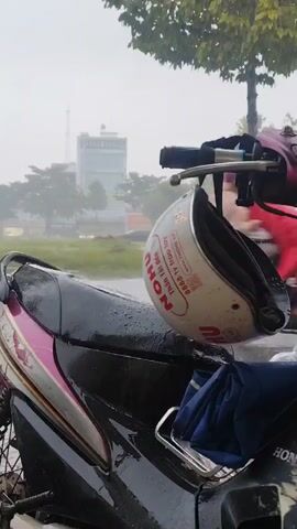 The video shows a close-up of a motorcycle parked outside during a rainstorm. A white helmet with red lettering is hanging from the handlebars. The seat of the motorcycle is black with purple and silver accents, and it is wet from the rain. A folded blue cloth is placed on the rear rack of the motorcycle. In the background, there is a blurred view of trees and a building, with rain visible in the air. The overall atmosphere is damp and overcast.