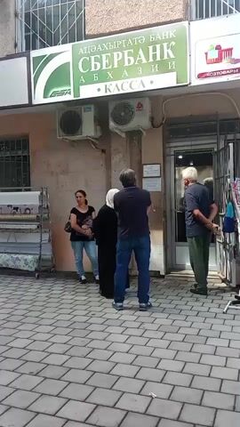 The video begins at eye level, showing a street scene outside a building with two storefronts. The left storefront displays a green sign with white lettering that reads "СБЕРБАНК" and "АБХАЗИИ," indicating a Sberbank branch. Below this sign, there are two air conditioning units mounted on the wall. To the right of the Sberbank is another storefront with a sign that reads "У РОЗЫ" and "ДЛЯ ДОМА, ШКОЛЫ И СВЯЗИ." This store appears to sell household goods, stationery, toys, electronics, and accessories.

In front of the Sberbank, a woman in a dark dress and headscarf stands with her back to the camera, facing the entrance. To her left, a woman in a blue shirt and jeans stands and looks towards the bank. A man in a dark blue shirt and jeans stands between the two women, also facing the bank.

In front of the "У РОЗЫ" store, a man with white hair wearing a light blue patterned shirt and a straw hat stands facing the entrance. He is talking to a woman with white hair wearing a dark patterned shirt and a green skirt. She is facing away from the camera, with her hands clasped behind her back.

The ground is paved with interlocking grey and beige bricks. Various items are displayed outside the "У РОЗЫ" store, including a rack of colorful balls hanging from the awning, shelves with boxes and other merchandise, and shopping bags. An open doorway to the "У РОЗЫ" store is visible, and inside, several people can be seen. The overall impression is a typical street scene in a town, with people queuing or shopping.