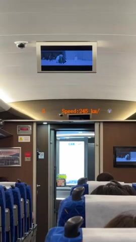 The video shows the interior of a train car, possibly a high-speed train. The camera first faces forward, showing a display screen indicating a speed of 245 km/h. The train has blue seats with headrests and wooden-paneled walls. There's a monitor displaying a movie or show above and in the background. Then the camera quickly pans to the window, showing a blurred view of the landscape passing by. The window appears to be wet or dirty, obscuring the view. The landscape appears to be flat and green.