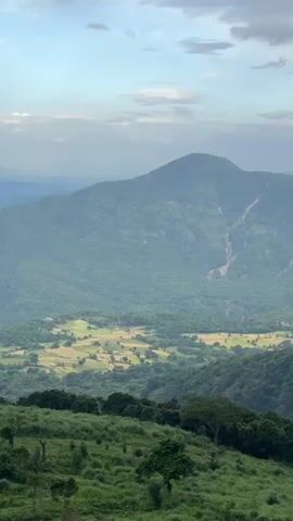 The video displays a panoramic view of a lush, mountainous landscape under a cloudy sky. In the foreground, dense green vegetation covers the hills, while in the midground, a valley interspersed with fields is visible. The background features several mountains, some with rocky cliffs, shrouded in a slight haze. The camera pans slowly to the right, revealing more of the mountain range. No people or animals are visible in the shot.