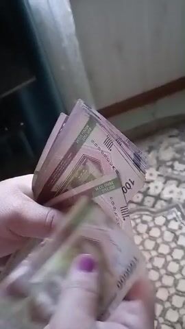 The video shows a close-up of a person's hands counting a fan of banknotes. The bills are predominantly light purple and green, with visible denominations of "50000" and "10000" in bold, dark lettering. There are also some numbers and symbols visible on the bills. The person has painted fingernails, with a vibrant purple polish on one finger. The background is slightly blurred, but appears to be an indoor setting with patterned flooring and a hint of furniture. The lighting suggests it could be daytime. The activity is the counting of money, implying a transaction or personal financial management. The focus is entirely on the hands and the currency, with no other subjects or clear environmental details to discern. The visible text confirms the currency denominations.