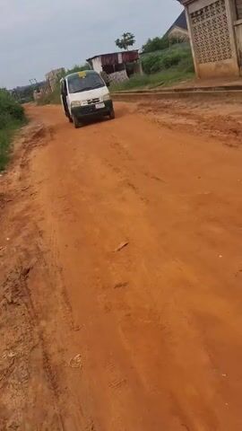 The video shows a white and green van driving on a reddish-brown dirt road in what appears to be a rural area. The road is uneven and marked with tire tracks. The van stops, and the sliding door on the side opens. The van then continues to drive, moving out of the frame. The surrounding environment consists of vegetation, a partially visible building with a distinctive pattern, and other distant structures. The sky is overcast.