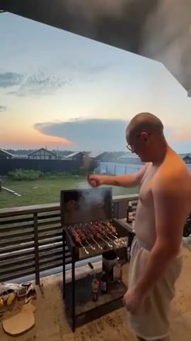 A man stands on a porch, grilling skewers of meat over coals in a metal grill. He wears glasses and light-colored shorts. The sky behind him is a dusk sky with clouds and a hint of sunset colors. The porch has a railing, and beyond that is a grassy yard with houses and fences. Smoke rises from the grill. There are bottles and a bucket on a shelf beneath the grill.