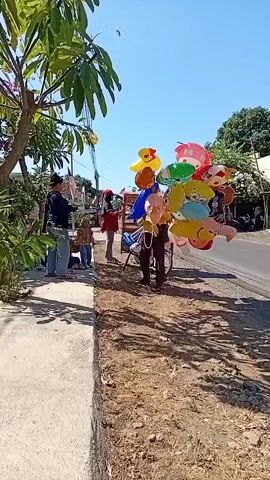 A street vendor pushes a bicycle laden with a large bunch of colorful, inflated balloons. The vendor is wearing dark clothing and is visible from the waist down, pushing the bike forward. To the left of the road, a sidewalk runs parallel to a grassy area. On the sidewalk, a person wearing a dark top and light blue jeans stands with their back to the camera, looking towards the vendor. A child in a light-colored top and dark pants stands to the right of this person, also looking towards the vendor. The balloons are various shapes and colors, including some cartoon character designs. The scene takes place outdoors under a clear blue sky, with trees and buildings visible in the background. The ground next to the road is dry and covered in dirt and sparse grass.