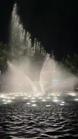The video shows a fountain show at night. Water shoots up from several points in a circular pool, illuminated by white lights underneath. The water forms various shapes, including tall columns and wide fan-like sprays. The background is dark, with some trees visible on the edge of the frame. In the distance, there are some blurred lights from nearby buildings.