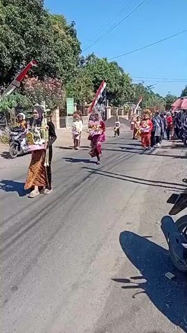 A parade is taking place on a paved road. The road is lined with trees and buildings. Indonesian flags are visible, mounted on poles along the side of the road. Several people are participating in the parade, wearing traditional clothing and costumes. Some individuals are carrying decorative items, such as bouquets of flowers. Spectators are gathered on the sides of the road, observing the procession. There are also motorcycles parked and moving in the vicinity. The sky is clear and blue, indicating a sunny day. The overall atmosphere appears festive.