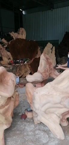 A worker wearing a face mask is sanding a large wooden sculpture inside a spacious workshop. The sculpture is made of light-colored wood with natural patterns and is surrounded by other wooden pieces, some finished and some raw. The workshop has a metal roof and walls, and various tools and equipment are scattered around, including a sanding machine, sandpaper, a fan, and a small cart. The worker moves around the sculpture, carefully sanding different sections.