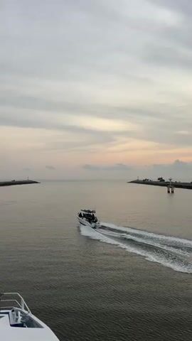 The video shows a motorboat moving away from the camera through the water. The boat is white with a dark top, and people can be seen on board. The boat leaves a white wake behind it. The water is calm and gray, and the sky is overcast with light pink and gray clouds. Two small islands with white structures on them are visible in the distance, marking the entrance to the waterway. Part of the white deck of a larger boat is visible in the foreground at the bottom of the screen. A speaker's voice can be heard in the background.