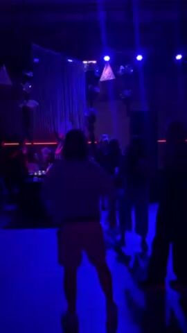 The video shows people dancing in a nightclub bathed in blue light.  The view is from behind a person wearing a white t-shirt and light-colored shorts.  Other figures are visible on the dance floor, as well as a DJ behind a booth with a sign that reads "Retro Mountain". There are stage lights and triangular decorations overhead.