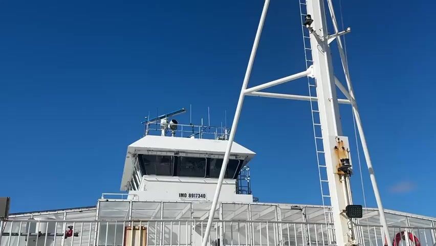 This media file captures a low-angle view of a white ferry against a clear, bright blue sky. The ferry's structure, including its bridge and a prominent mast with a ladder, dominates the frame. The bridge is a multi-paned cabin, likely housing the controls for navigating the vessel. Above the bridge, various antennae and a radar scanner are visible, suggesting the ferry is equipped for maritime operations. The ferry's hull is partially visible, featuring white railings and what appears to be a section of enclosed decking.

The scene is set in an open environment, indicated by the expansive sky and the nautical elements. The time of day appears to be daytime, with bright sunlight casting shadows and illuminating the white surfaces of the ferry. The weather seems to be fair, with no clouds disrupting the deep blue of the sky. The location is implied to be coastal or a waterway, given the presence of a ferry.

Notable details include the vessel's identification number, "IMO 8917340," clearly displayed on the side of the bridge. There are also small lights and possibly security cameras mounted on the mast. A red life preserver is visible on the right side of the ferry's railing, a standard safety feature. A person in a red jacket is visible on the left side of the ferry, adding a human element to the otherwise structural composition. The overall mood is one of calm and clarity, conveyed by the clean lines of the ferry and the unblemished sky. The image's focus is on the functional aspects of the ferry's design against a backdrop of natural elements. The location context of Korsnes, Norway, suggests this ferry operates in Norwegian waters.