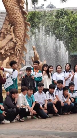 A group of about 20 teenagers is posing for a group photo in an outdoor area. They are arranged in a semi-circle, some crouching and some standing. A young woman with a megaphone appears to be directing them. In the background, there is a large, golden dragon statue with water spraying behind it. Some of the teenagers are waving. The group consists of both males and females, dressed in a variety of casual clothing and school uniforms. The area is paved and there are trees and buildings visible in the background. The overall impression is that the group is enjoying a photo opportunity.