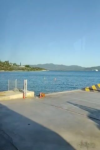 The video shows a view from a dock on a sunny day. The water is calm and blue, with some small waves. In the background, there are green hills covered with trees and some houses. There are a few people swimming in the water near the shore. On the dock, there are a few signs. One sign is yellow and has a logo that says "Kaoil" and a distance of 650m. Another sign is green and points to the left, with text that says "Athens", "Thessaloniki", and an arrow. There is also a blue sign with a white "P" on it, indicating parking, and the word "TAXI". A small blue boat is parked on the dock. The overall impression is a peaceful seaside scene, possibly a small port or beach town.