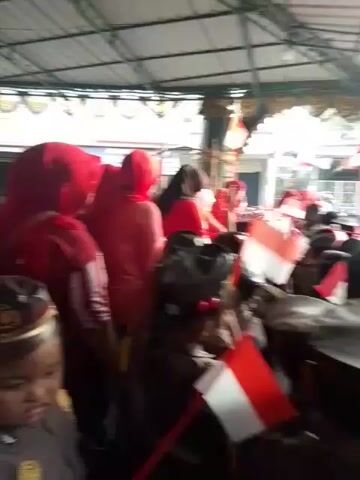 The video shows a crowd of people, many of whom are children dressed in uniforms resembling police or military attire. They are wearing hats and dark-colored uniforms adorned with patches and medals. Some of the children are holding small Indonesian flags, which are red and white. The scene appears to be indoors, possibly under a covered structure with a metal roof. The lighting is somewhat dim, with some bright areas suggesting natural light from an opening. The children seem to be gathered together, possibly for an event or ceremony. One child in the foreground, wearing a police-like uniform and hat, is seen rubbing their eye with their finger. The overall atmosphere seems to be one of participation in a patriotic or ceremonial event.