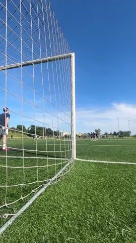 The video shows a man practicing soccer on a sunny day. The camera is positioned behind the soccer goal, looking out onto the field. The man, wearing a dark shirt and light shorts, stands to the left of the goal. He kicks a soccer ball, sending it into the upper left corner of the net. He then walks off the field toward the right. Other people are scattered on the field in the background, practicing soccer as well. The sky is blue with some wispy clouds. The field is green and well-maintained.