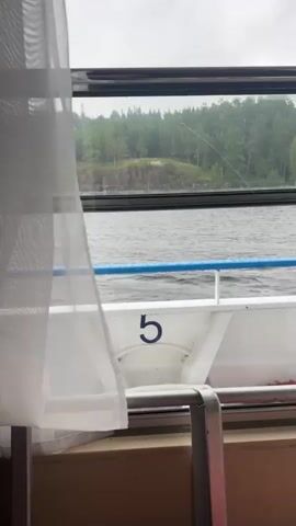 The video shows a view from inside a boat looking out a window. A sheer white curtain is in the foreground on the left. The window shows a body of water with small waves. In the distance, there is a small, rocky island covered in trees. The sky is overcast. Part of the boat is visible in the foreground, including a white railing with a blue stripe and the number 5 painted on a white surface. There is also a white plastic container and a coiled red rope.