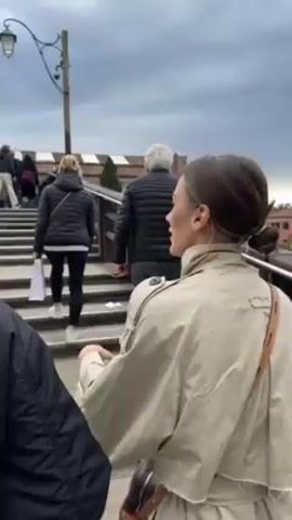 The video shows a group of people walking up a set of stairs on a bridge, presumably in Venice, Italy. The focus is on a woman in a tan trench coat who is gesturing and talking. Other people are ahead of her on the stairs, including a woman in a black puffer jacket and leggings, and a man in a similar black jacket and jeans. The sky is overcast. The railing of the bridge is wooden, and you can see colorful buildings in the background.