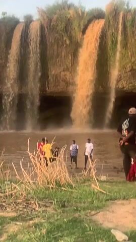 The scene shows a waterfall with brownish water cascading over a rocky ledge into a river. Several people are standing in the water near the base of the waterfall, while others are sitting or standing on the grassy bank. The people are dressed in various casual clothes. The sky is light blue and the lighting suggests it is daytime. Some dry vegetation is visible in the foreground.
