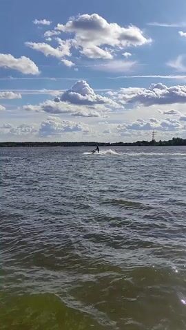A person is riding a jet ski on a body of water. The water is choppy and a wake is visible behind the jet ski. The sky is blue with white clouds. A distant shoreline with trees is visible. The overall scene is one of recreation on a sunny day.