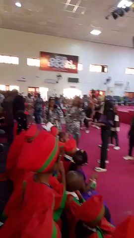 The video shows a group of people, including children and adults, in what appears to be an indoor event space with a pink carpeted floor and a white ceiling. A banner hangs from the ceiling displaying "2026 OPEN DOORS I'm A Winner". Many of the children are wearing red outfits with green trim, and some of the adults are wearing patterned clothing. Several people are seen dancing or moving to music. The lighting is bright, suggesting it is daytime or the space is well-lit. The overall atmosphere appears to be celebratory.