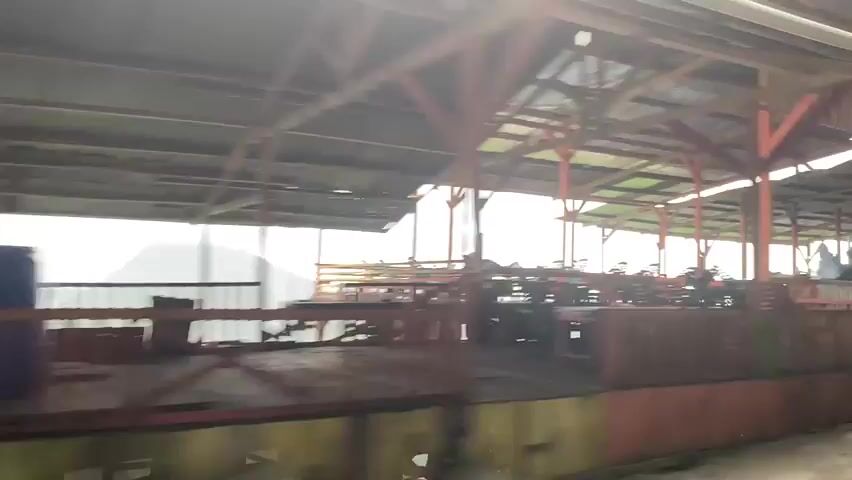 The video shows an outdoor dining area with several tables and chairs set up under covered roofs. People are seated at some of the tables. There are also several vehicles present, including cars and motorcycles, parked in the vicinity. The environment appears to be a scenic location with a view of a body of water or a valley in the background, under an overcast sky. The structures are made of wood and metal, with corrugated roofs. The scene suggests a casual, possibly tourist-oriented establishment.