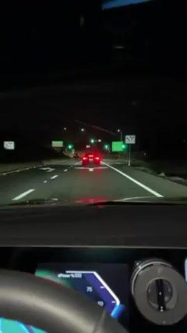 A stationary, first-person view from inside a car stopped at a traffic light at night. The dashboard is visible, including a digital display showing an energy level indicator, a gear selector knob, and a phone mounted to the right. Through the windshield, a red car is ahead, stopped at the same light. The surroundings include streetlights, highway signs, and multiple arrows directing lane use. The traffic light changes to green.