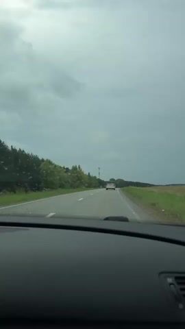 This video shows a view from the inside of a car looking out at a road. The sky is overcast with dark clouds. On the left side of the road, there are trees and bushes. On the right side of the road, there is a field of grass. A red car is driving in front of the car from which the video is being recorded. The road appears to be in a rural area. The video is short and does not contain any significant actions or interactions.