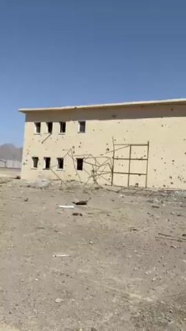 The video shows a damaged two-story building, possibly in a war zone. The building is beige with many bullet holes and structural damage. The windows are missing. In front of the building, there is a severely damaged and burned-out station wagon. Debris litters the ground. The surrounding terrain appears arid and desolate. The sky is clear and blue. A rudimentary metal structure stands near the building. The camera pans across the scene, revealing the extent of the damage. There are no people visible.