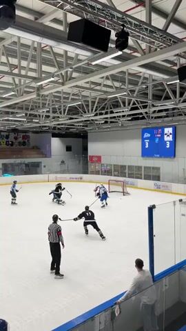 The video shows an ice hockey game. Several players in uniforms are on the ice, some with sticks. A referee in a black and white striped shirt is also on the ice. There is a net and a scoreboard visible in the background. The scoreboard shows the time as 11:35 and the score as 3-3. The arena appears to be indoors with a high ceiling and exposed beams. There are spectators in the foreground.