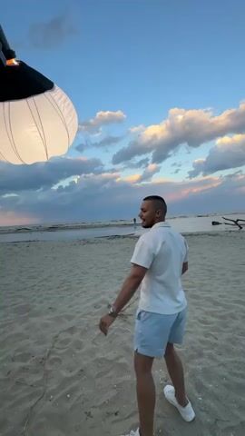 A man in a white polo shirt and blue shorts is on a beach, looking toward the sea where another person is standing. The sky is blue with white and purple clouds. A large, round, white light is suspended overhead, attached to a black stand. A second man, wearing a green t-shirt and black cap, is standing behind the light stand, looking at the camera in his hands. In the distance, there are other people on the beach and a piece of driftwood in the water. The beach is sandy.