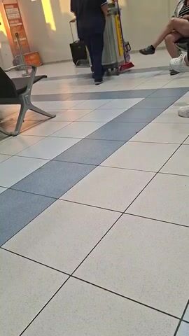 The video shows a tiled floor in what appears to be an airport or train station waiting area. People are seen walking and sitting in the background. In the foreground, the feet of several individuals are visible, some wearing flip-flops and others wearing sneakers. The lighting suggests it is daytime, with sunlight casting shadows on the floor.
