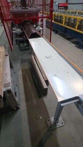 The video shows an automated machine shaping metal sheets into a curved form. A brown, curved metal sheet is seen emerging from the machine onto a white metal conveyor belt, which is inclined at an angle. The machine is enclosed in a red protective cage. A wooden pallet and several wooden planks are positioned near the machine's exit. The machine is in an industrial setting.