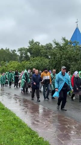 A group of people, many wearing brightly colored raincoats, are walking in a procession down a wet asphalt path lined with grass. The sky is overcast and the ground appears to have been recently rained on, with puddles visible. In the background, there are trees, a building with a blue roof, and a lamppost. Some of the individuals are looking up or to the side, while others are engaged in conversation. The overall atmosphere appears to be one of a planned walk or pilgrimage, possibly in adverse weather conditions.