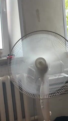 The video shows a close-up of a white electric fan spinning rapidly, creating a blurred effect with its blades. The fan is positioned in front of a window, with a radiator visible on the left and a textured wall in the background. The lighting suggests it is daytime, with natural light entering from the window. There are no people or animals visible in the frame. The fan appears to be the sole focus of the video.