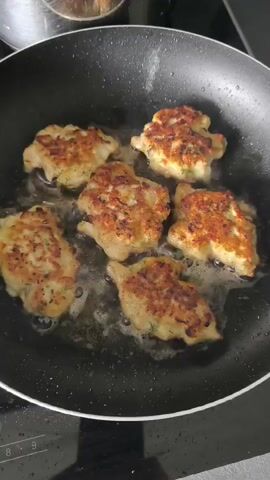 Six chicken cutlets are frying in oil in a black non-stick frying pan on a stovetop. The cutlets are golden brown and appear to be seasoned with herbs. The pan is on a black glass stovetop with visible control markings.