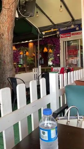 A medium shot shows a woman sitting at an outdoor cafe table, talking on a mobile phone held to her ear. She has dark hair and is wearing a white shirt. In front of her on the dark brown table are a glass with a green drink and a purple straw, a white coffee cup and saucer, and a silver French press. To her left, also on the table, are a clear plastic water bottle with a blue cap, an ashtray, a pack of Marlboro cigarettes, and a glass vase with colorful candy-like stones at the bottom holding artificial flowers. A white picket fence runs across the mid-ground. In the background, other patrons are seated at tables under an awning with string lights, and signs for "MERKEZ BÜFE" and "DONDURMA" are visible. The woman occasionally gestures with her free hand as she speaks on the phone.