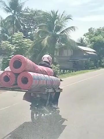 The video shows a person riding a motorcycle on a paved road. The motorcycle is heavily loaded with rolled-up mats secured on a platform behind the rider. The rider is wearing a helmet. The background features trees, buildings, and a clear sky. The camera appears to be moving, likely taken from another vehicle following the motorcycle.