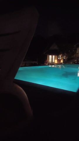At night, a person wearing shorts climbs over the edge of a swimming pool. The pool is lit with turquoise light. In the background are two small buildings with lit windows.