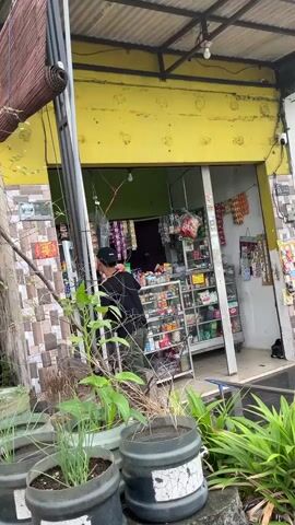 The video shows a person wearing a black hat and black shirt in a convenience store. The store has yellow exterior walls and is filled with shelves stocked with various products, including cigarettes, snacks, and drinks. Outside the store, there are potted plants and a tiled wall. The person appears to be browsing or selecting items from the shelves.