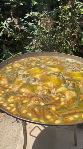 A large, wide paella pan filled with a simmering mixture of rice, green beans, and pieces of meat is positioned outdoors on a metal stand. The paella is actively bubbling, with steam rising from the surface. The background consists of lush green foliage, including rose bushes with red roses in bloom, and trees under a bright blue sky. The camera pans slightly, offering a closer view of the cooking paella and then a wider view of the surrounding garden.