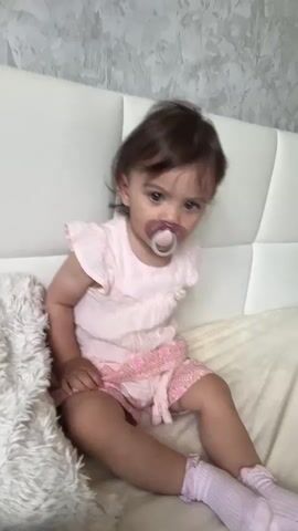 A young child with brown hair and a pacifier is sitting on a bed. The child is wearing a light pink dress and pink shorts with a lace trim. The child is also wearing pink socks. The bed has a white headboard. There is a fluffy white blanket on the left side of the bed. The background is a gray textured wall. The child blinks and moves their head slightly.