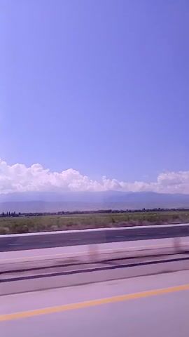 A side-view, from a moving vehicle, of a highway. The road in the foreground has a grey surface and a yellow line on the right. Beyond the road, there is a green field, then a line of trees and mountains in the background, all under a clear blue sky with a few white clouds.