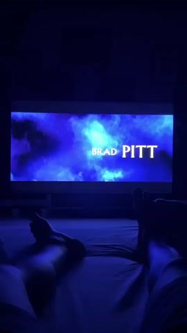 The video shows a screen displaying the name "BRAD PITT" in large, bold letters against a vibrant, smoky blue background. The text is illuminated, appearing as though it's glowing. Below the screen, a person's legs and feet are visible, stretched out on what appears to be a bed or sofa covered with a white sheet. The room is dimly lit, with the primary light source emanating from the screen, casting a strong blue hue over the scene and the person's legs. The overall ambiance is dark and relaxed, suggesting someone is watching a movie or TV show in their home. The time of day is likely evening or night due to the darkness.