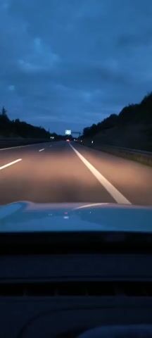 The video shows a first-person perspective of driving on a highway at dusk. The sky is dark blue with some clouds. The headlights of the car illuminate the road, which has white lane markings. There is a sign overhead in the distance. Trees and hills are visible on either side of the highway.