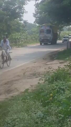 The video shows a truck driving down a road. The truck is white with a yellow tarp covering its cargo. There is a white car behind the truck. On the left side of the road, there is a person on a bicycle. The road is surrounded by trees and greenery. The sky is visible and appears to be hazy. The overall scene suggests a daytime outdoor setting, possibly in a rural or suburban area.