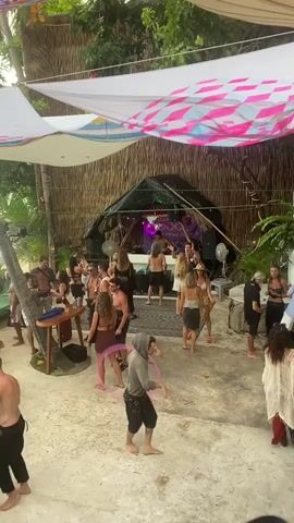 A party is happening in an outdoor space with a dirt floor. A DJ booth made of natural materials is set up under a roof of straw. A crowd of people in casual, resort-style clothes dances in front of the booth. A man in a gray hooded shirt and black shorts stands facing the camera, twirling a pink hula hoop around his arm. A pink-and-white tarp is stretched overhead to provide shade.